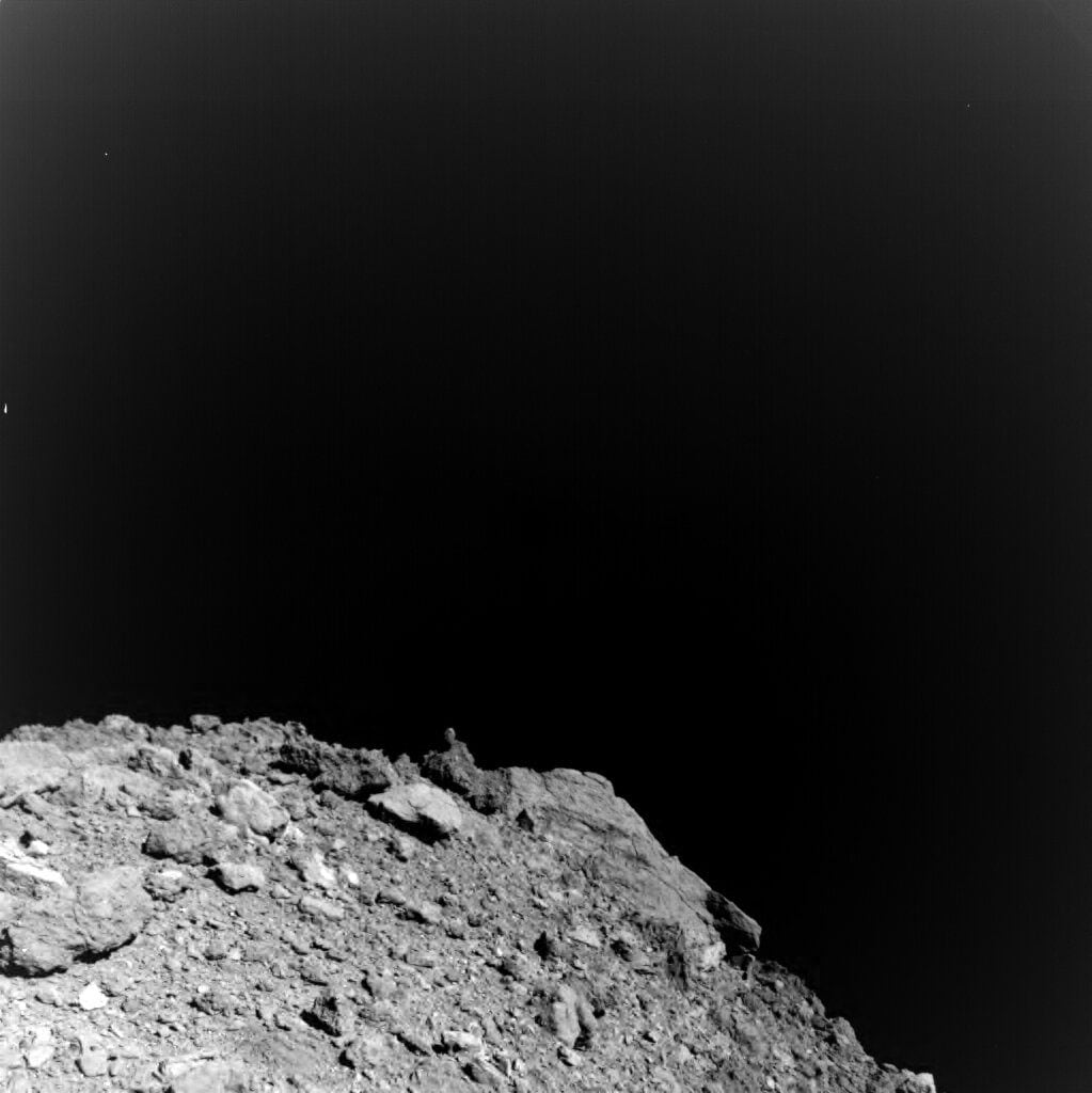 The first image acquired by the MASCAM camera system on Hayabusa2 during its descent, shortly after separation from the landing module at a height of 41 meters with a viewing direction towards the south pole. Particularly striking is a huge block near the South Pole, which stands out clearly above the horizon line named 'Otohime Saxum'. It is up ~100 meters tall. Image: Jaumann et al/Science