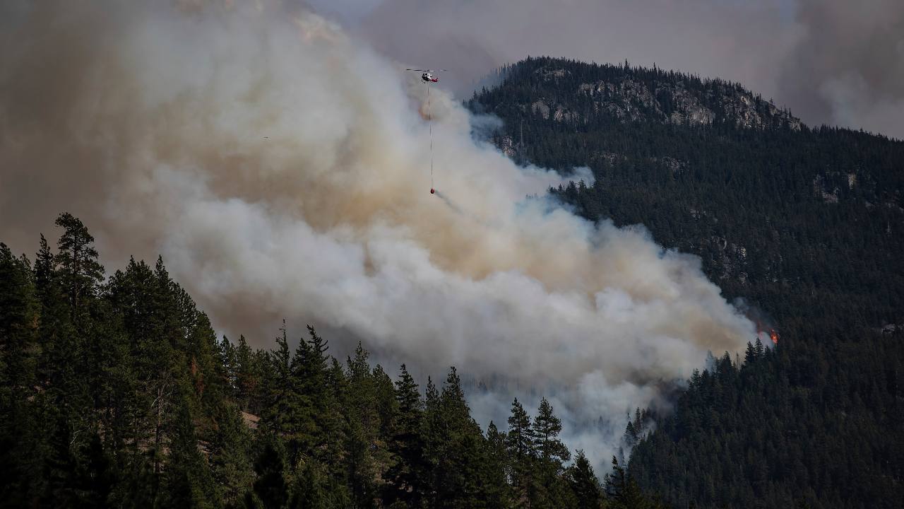 A helicopter carrying a water bucket flies past the Lytton Creek wildfire burning in the mountains near Lytton, British Columbia, on Sunday, Aug. 15, 2021. (Darryl Dyck/The Canadian Press via AP)