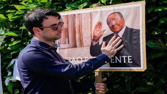 A man poses next to a poster of Silvio Berlusconi outside the latter’s residence. In the case of the latter, flags are flown at half-staff and all political events not involving charity are put on hold, but it is otherwise business as usual. AP