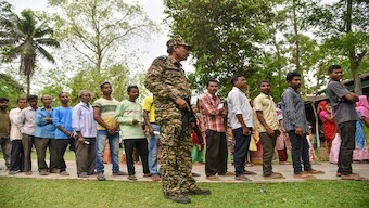 A security personnel stands guard as voters wait in a queue at a polling station to cast their votes for the first phase of Lok Sabha elections, in Nagaon district, 19 April 2024. PTI