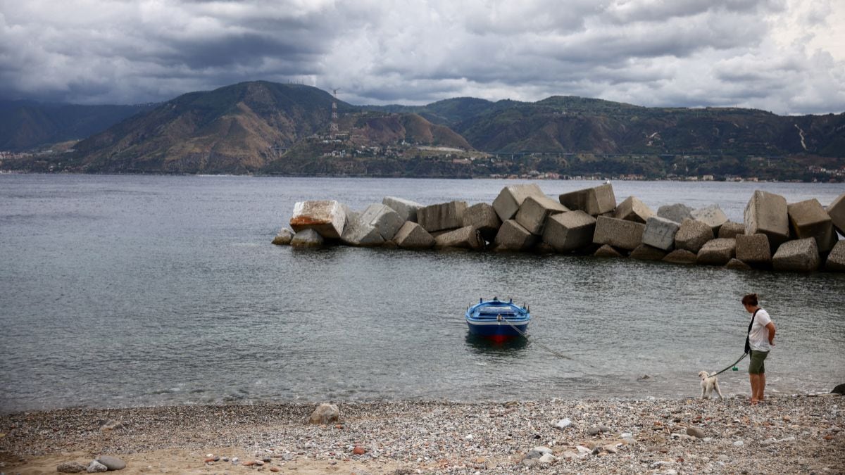 A woman walks with her dog along the Sicilian coast with the Calabrian shoreline visible across the Strait of Messina, Italy, August 5, 2025. The area is part of the planned site for a suspension bridge connecting Sicily to mainland Italy, with construction expected to begin in the second half of 2025. Reuters