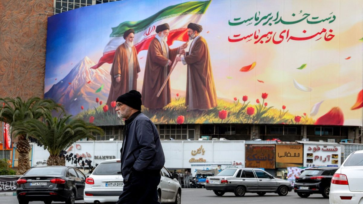 A man walks near a banner displayed at Valiasr Square in central Tehran, depicting Iran's late Supreme Leader Ayatollah Ruhollah Khomeini watching as his successor the late Ayatollah Ali Khamenei hands over a national flag to his son and new Supreme Leader Mojtaba Khamenei. (Photo: AFP) A man walks near a banner displayed at Valiasr Square in central Tehran, depicting Iran's late Supreme Leader Ayatollah Ruhollah Khomeini watching as his successor the late Ayatollah Ali Khamenei hands over a national flag to his son and new Supreme Leader Mojtaba Khamenei. (Photo: AFP)