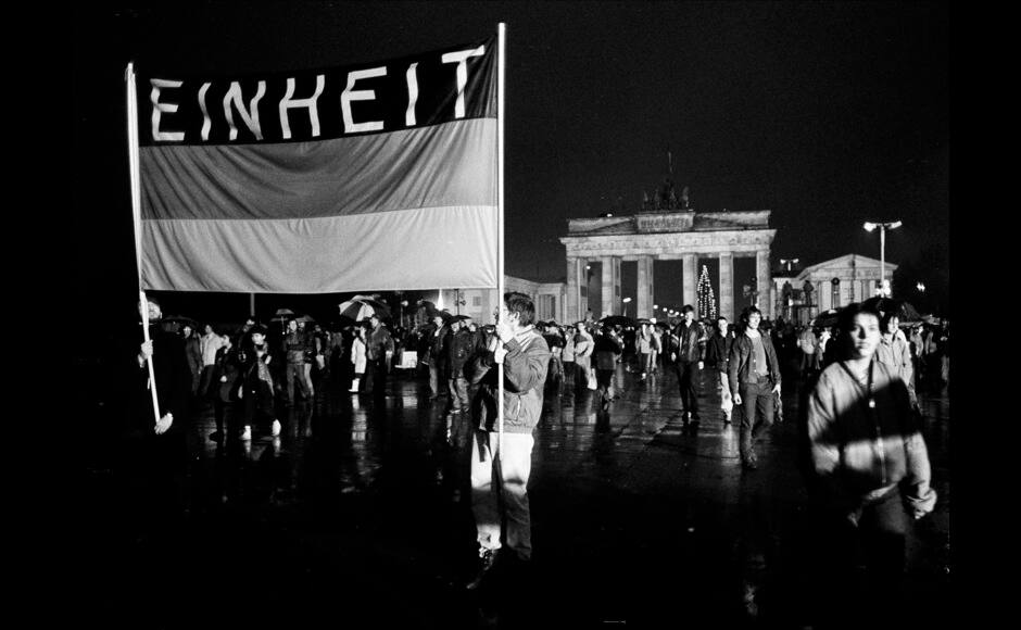 People hold a German flag with the word reading 'Unity' as they cross the newly opened checkpoint between East and West Berlin at the Brandenburg Gate, December 22, 1989. REUTERS/Fabrizio Bensch (GERMANY ANNIVERSARY POLITICS) - RTR25OX0