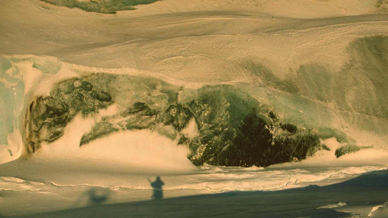 A close-up image of an iceberg near Mawson Station, from November, 1988. Much of it covered by snow, at the far left is bubbly blue-white glacier ice. The center of the picture shows the clear, dark green, bubble-free marine ice. The boundary between glacier ice and marine ice is nearly vertical. Image credit: Warren et al