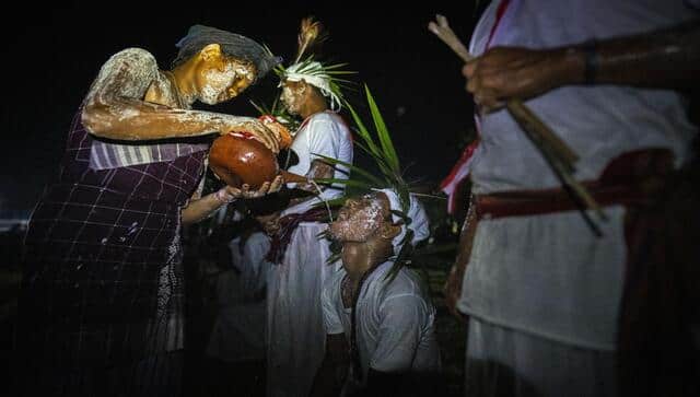An Indian Rabha tribal girl in traditional attire with face smeared with rice flour pours rice beer as holy water to a Hindu priest after he walks barefoot over burning charcoal as part of rituals during Baikho festival