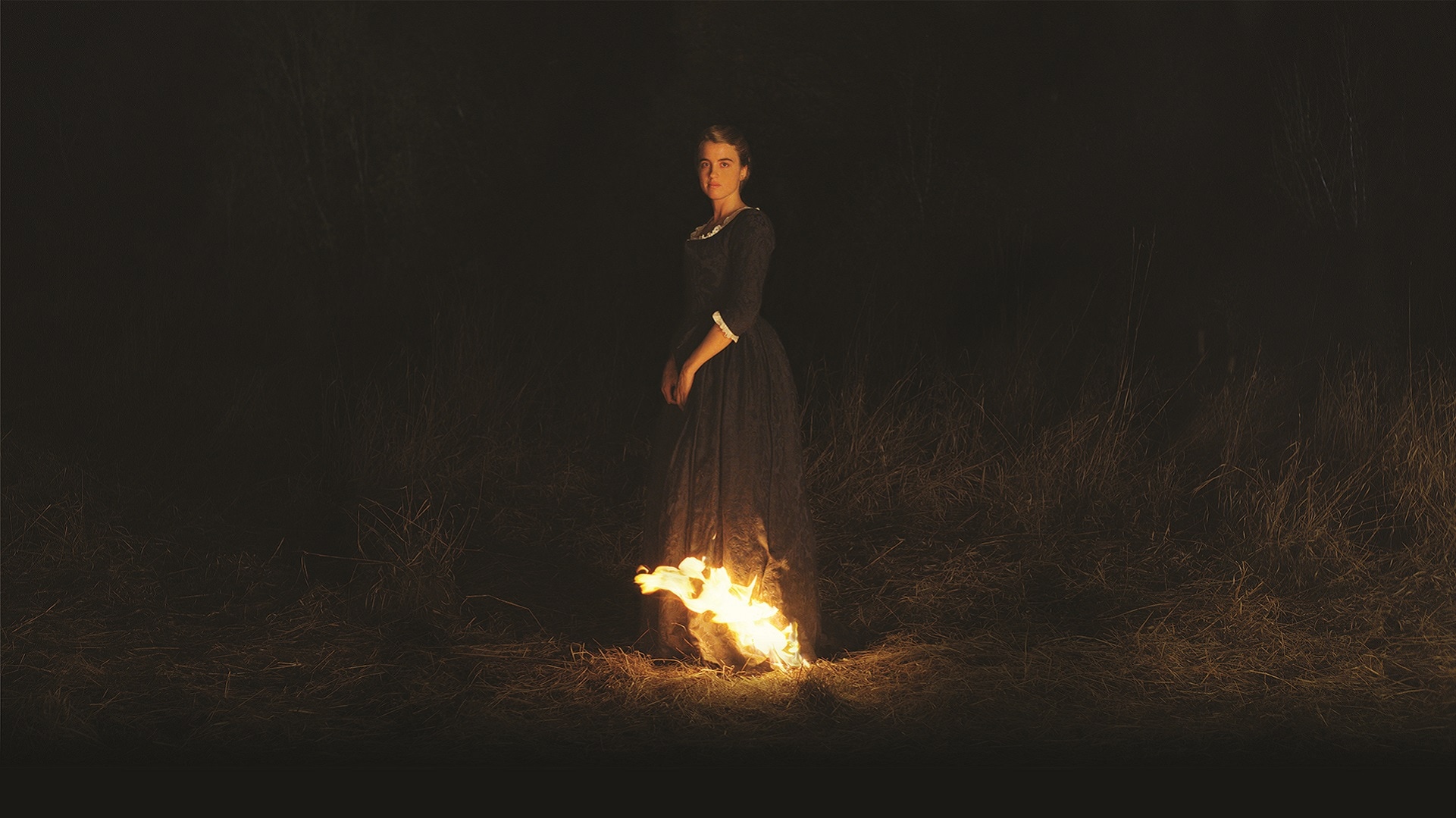 Image is from the film 'Portrait of a Lady on Fire' (2019). In almost complete darkness, a woman stands in the centre of the frame. The bottom on her dress is on fire.